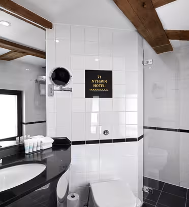 Modern bathroom with white and black tiled walls, a circular mirror, and a sleek black countertop with a sink. Towels and toiletries are neatly arranged. Wooden beams accent the ceiling. A sign reads 71 Nyhavn Hotel on the wall.