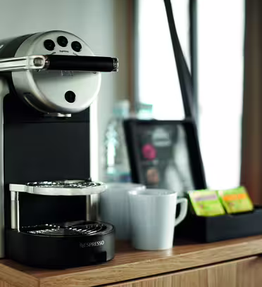 A sleek espresso machine sits on a wooden counter, accompanied by two white mugs, a set of coffee pods, and a water bottle in the background. It creates a cozy coffee nook setting.