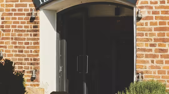 Entrance of Ristorante Il Rosmarino with a brick facade and a black sign. Outdoor table with chairs, glassware, and potted plants in the foreground, suggesting a relaxed dining atmosphere.