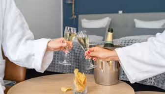 Two people in white robes clinking champagne glasses in a hotel room. A bottle of champagne sits in an ice bucket on a wooden table with a glass of potato chips in the foreground. A neatly made bed is visible in the background.
