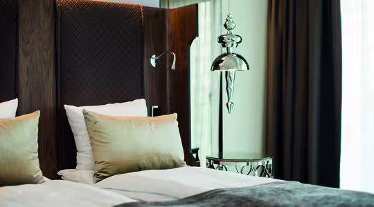 A modern hotel room features a bed with white linens and beige pillows. The headboard is textured with a diamond pattern. A sleek silver lamp hangs above a round table, and a large window with dark curtains provides natural light.